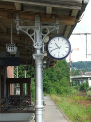 Gleise Eine Bahnhofsuhr aus Metall zeigt 8:57 Uhr, im Hintergrund grünes üppiges Buschwerk.