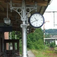 Gleise Eine Bahnhofsuhr aus Metall zeigt 8:57 Uhr, im Hintergrund grünes üppiges Buschwerk.