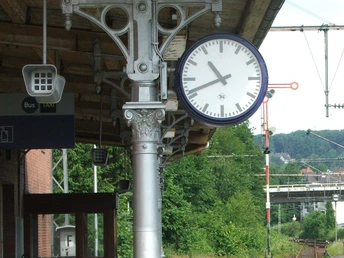 Gleise Eine Bahnhofsuhr aus Metall zeigt 8:57 Uhr, im Hintergrund grünes üppiges Buschwerk.