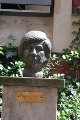 Büste von Clara Ernst aus Bronze auf einem Sockel, umgeben von Efeu und Ziegelsteinmauer.
