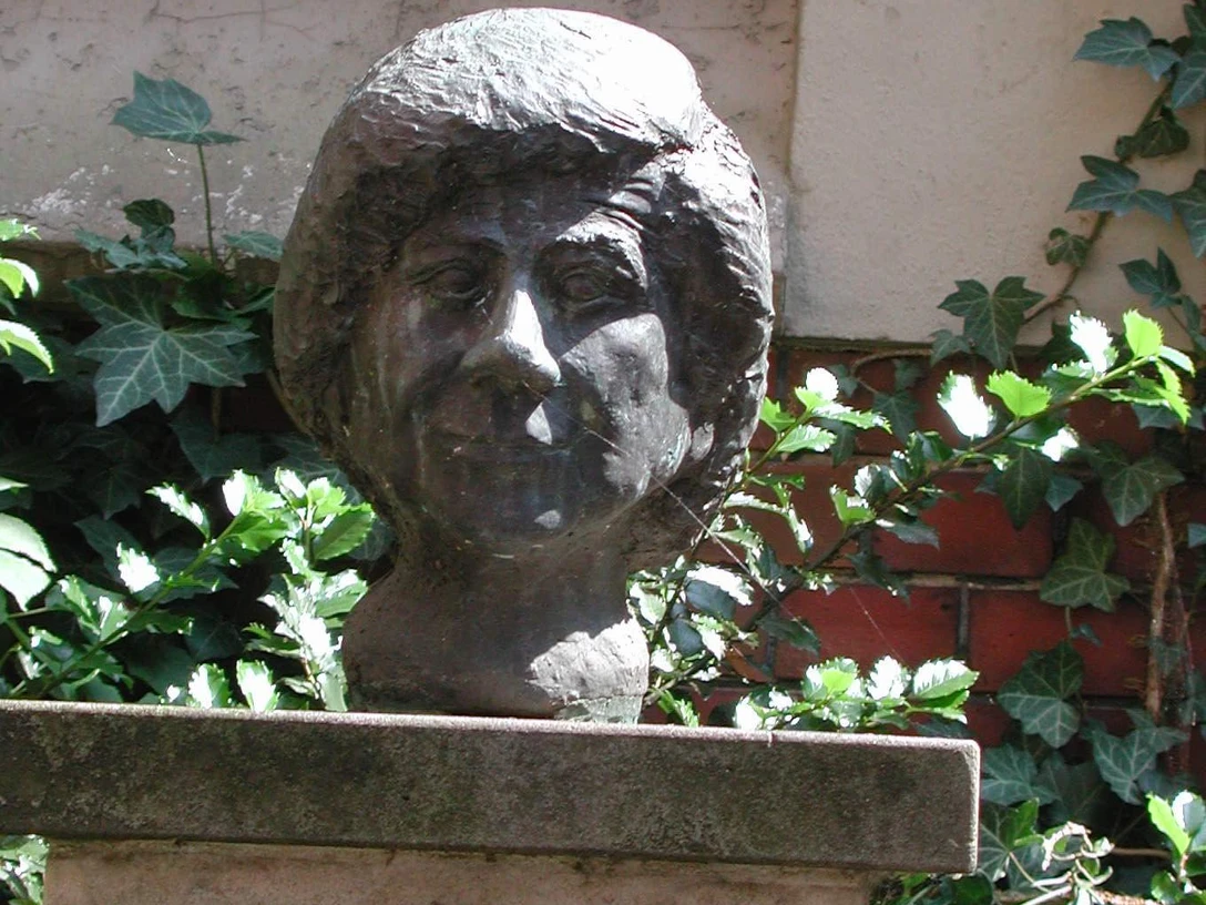 Büste von Clara Ernst aus Bronze auf einem Sockel, umgeben von Efeu und Ziegelsteinmauer.