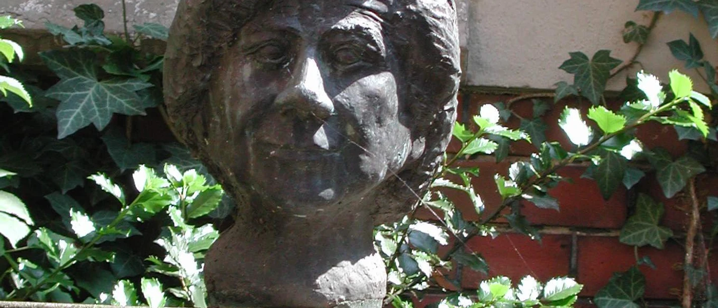 Clara-Ernst-Büste Büste von Clara Ernst aus Bronze auf einem Sockel, umgeben von Efeu und Ziegelsteinmauer.