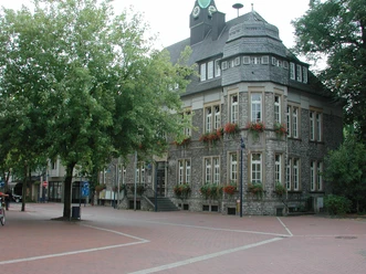 Historisches Rathaus mit grauer Steinfront, umgeben von Bäumen, roten Blumenkästen an den Fenstern.