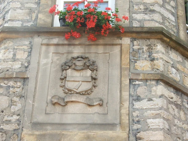 Ein steinernes Stadtwappen ziert die Fassade des Rathauses, umrahmt von roten Geranienblüten.