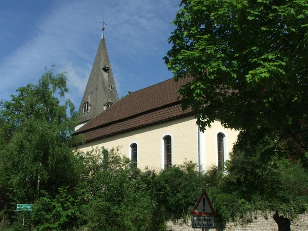 Die Kirche Stapelage mit spitzem Turm, umgeben von Bäumen und strahlend im Sonnenlicht.