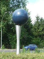 Kunstwerk: Metallene Kugel auf konischem Standfuß, umgeben von Grün und vorbeifahrendem Auto.