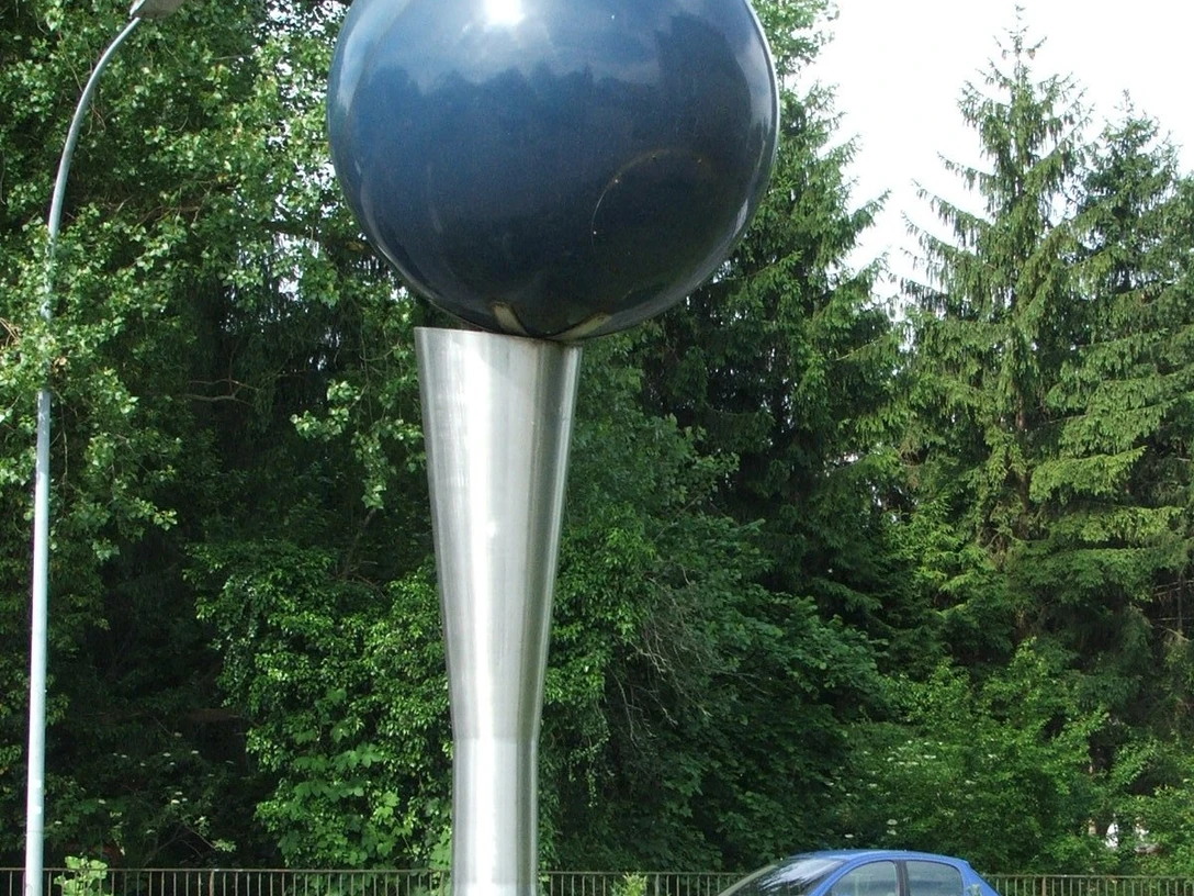 Kunstwerk: Metallene Kugel auf konischem Standfuß, umgeben von Grün und vorbeifahrendem Auto.