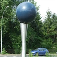 Volksbankkugel Kunstwerk: Metallene Kugel auf konischem Standfuß, umgeben von Grün und vorbeifahrendem Auto.