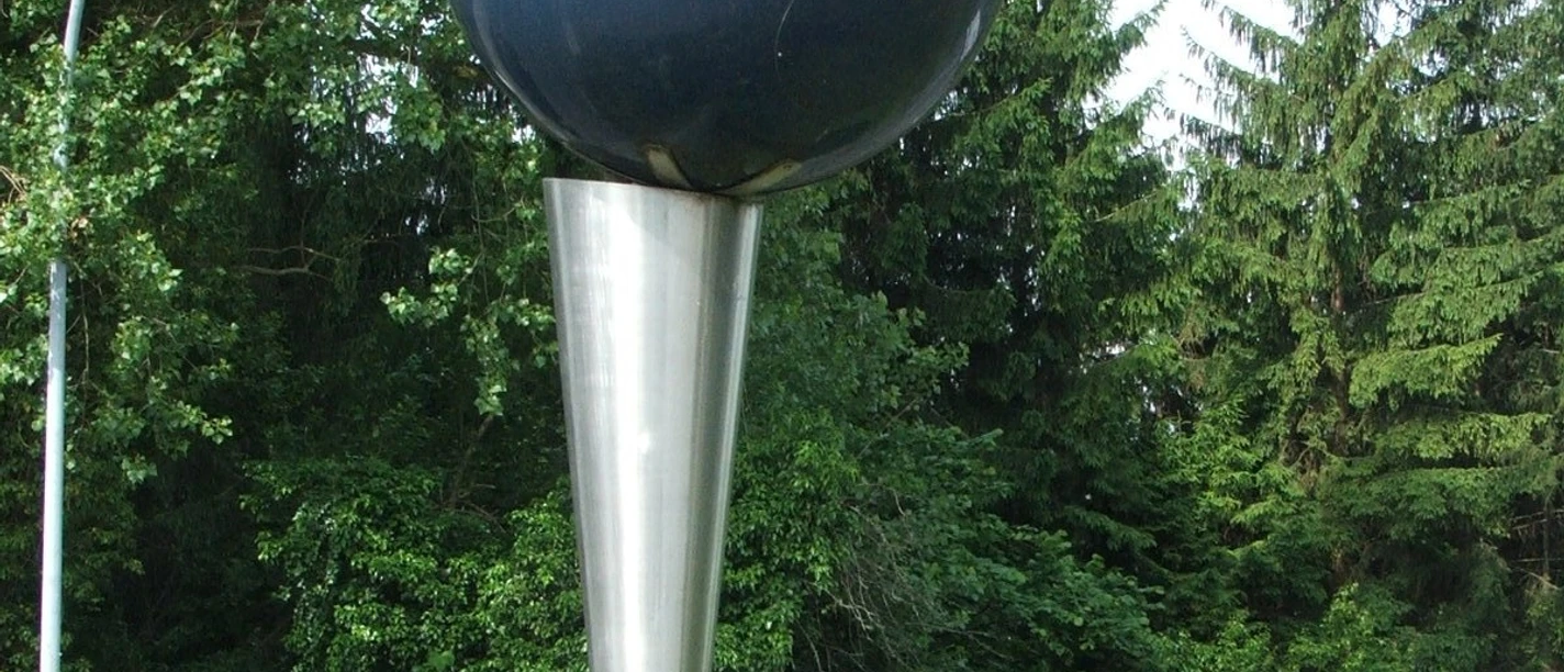 Volksbankkugel Kunstwerk: Metallene Kugel auf konischem Standfuß, umgeben von Grün und vorbeifahrendem Auto.