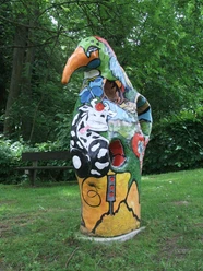 Schampus Fiaskus Farbenfrohe, fantasievolle Skulptur eines Vogels in einem grünen Parkumfeld, umgeben von Bäumen.