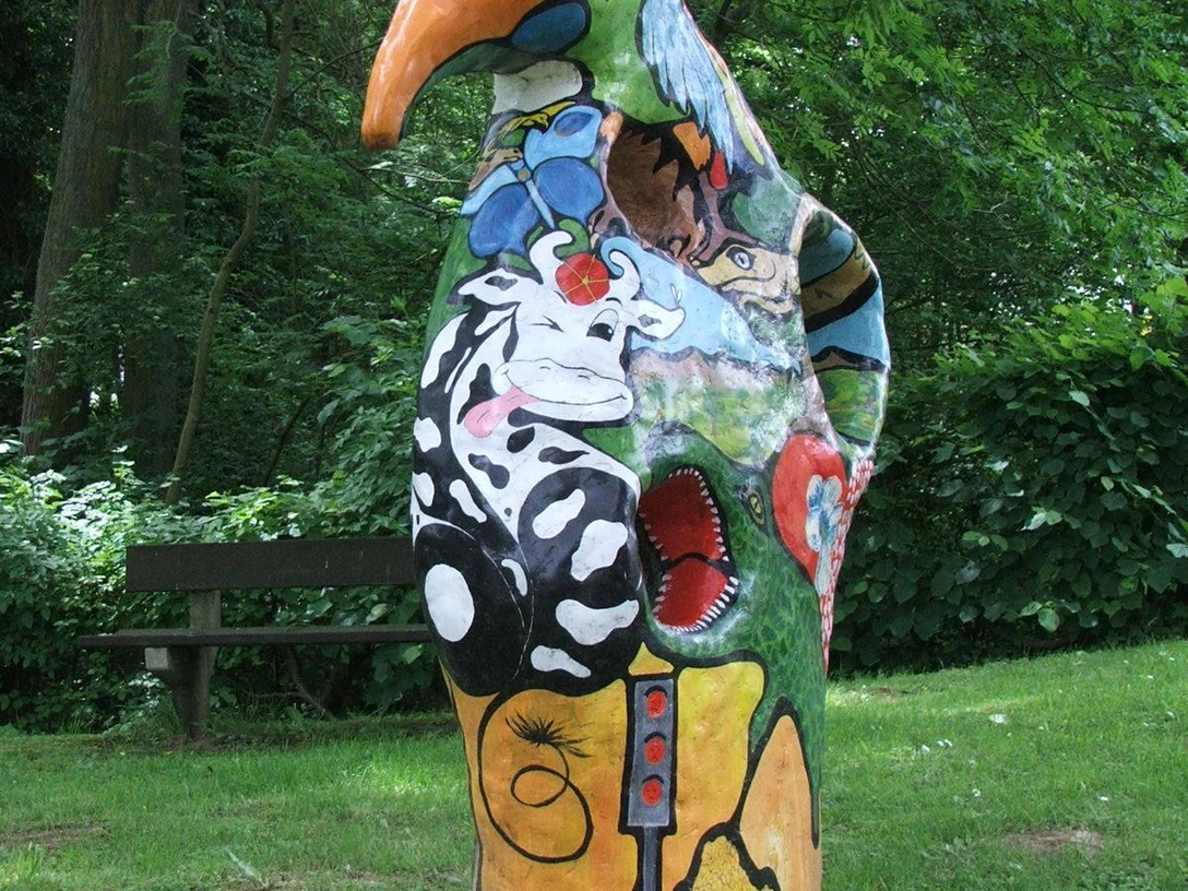 Schampus Fiaskus Farbenfrohe, fantasievolle Skulptur eines Vogels in einem grünen Parkumfeld, umgeben von Bäumen.