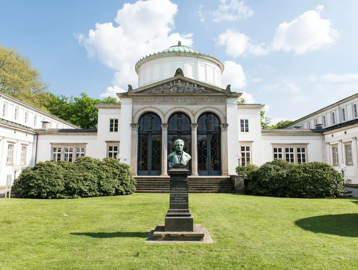 Auf dem Bild ist das Badehaus I mit seiner weißen Fassade und dem majestätischen Eingang zu sehen. Vor dem Gebäude befinden sich zwei Büsche und ein gepflegter Vorgarten. Im Vordergrund ist eine Skulptur zu erkennen, die das Bild zusätzlich ziert.