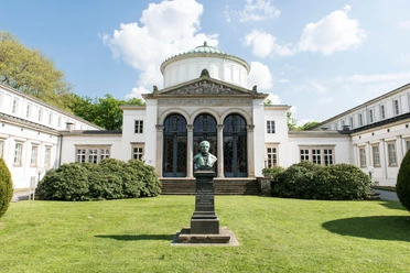 Badehaus I Auf dem Bild ist das Badehaus I mit seiner weißen Fassade und dem majestätischen Eingang zu sehen. Vor dem Gebäude befinden sich zwei Büsche und ein gepflegter Vorgarten. Im Vordergrund ist eine Skulptur zu erkennen, die das Bild zusätzlich ziert.