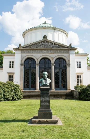 Badehaus I Auf dem Bild ist das Badehaus I mit seiner weißen Fassade und dem majestätischen Eingang zu sehen. Vor dem Gebäude befinden sich zwei Büsche und ein gepflegter Vorgarten. Im Vordergrund ist eine Skulptur zu erkennen, die das Bild zusätzlich ziert.