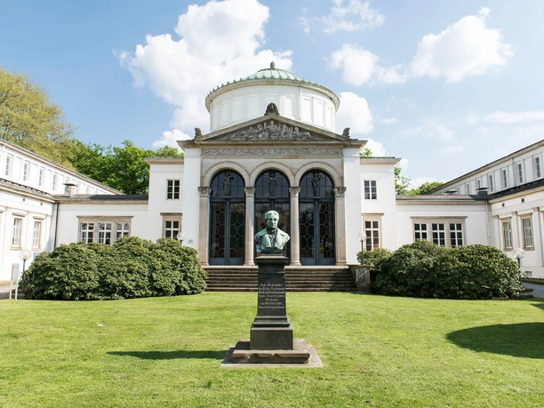 Badehaus I Auf dem Bild ist das Badehaus I mit seiner weißen Fassade und dem majestätischen Eingang zu sehen. Vor dem Gebäude befinden sich zwei Büsche und ein gepflegter Vorgarten. Im Vordergrund ist eine Skulptur zu erkennen, die das Bild zusätzlich ziert.