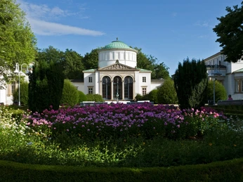 Neoklassizistisches Badehaus mit grüner Kuppel, umgeben von blühenden Gärten und gepflegten Hecken.