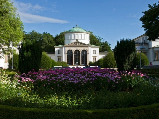 Neoklassizistisches Badehaus mit grüner Kuppel, umgeben von blühenden Gärten und gepflegten Hecken.