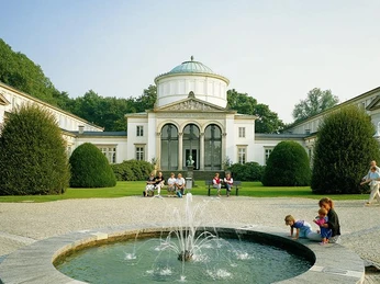 Historisches Badehaus mit weißer Fassade, Springbrunnen im Vordergrund, umgeben von Grünflächen.