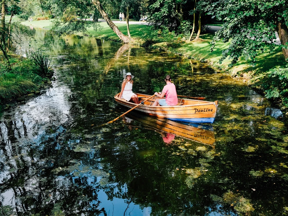 Rudern im Kurparksee im Landschaftsgarten Zwei Personen rudern in einem klassischen Holzboot auf einem von Bäumen gesäumten Kurparksee.