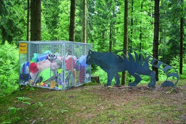 Mensch und Natur Ein Metallkäfig voller bunter Müllsäcke im Wald, daneben eine Tierfigur aus Metall.