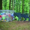 Mensch und Natur Ein Metallkäfig voller bunter Müllsäcke im Wald, daneben eine Tierfigur aus Metall.