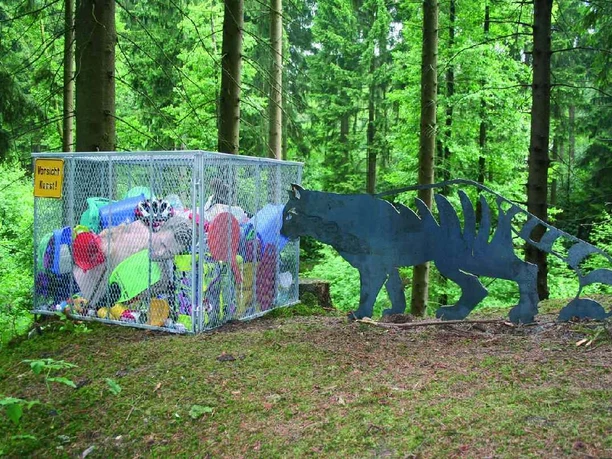 Mensch und Natur Ein Metallkäfig voller bunter Müllsäcke im Wald, daneben eine Tierfigur aus Metall.