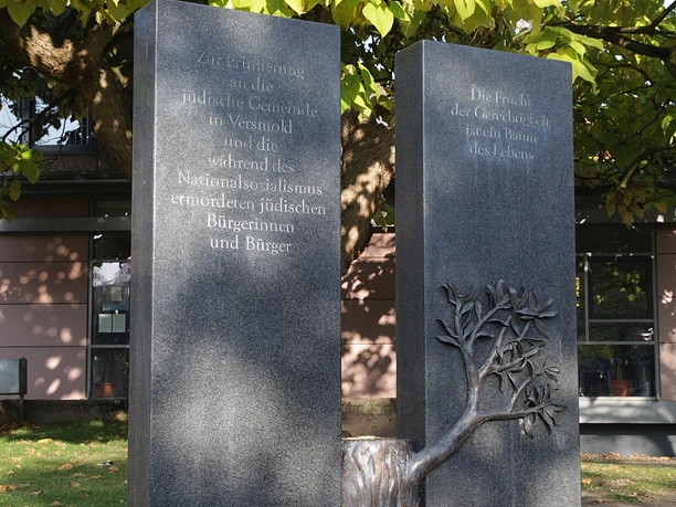 Erinnerungszeichen vor dem Rathaus Zwei dunkle Gedenksteine mit Gravuren, umrahmt von einem Baum, symbolisieren Erinnerung an Opfer.