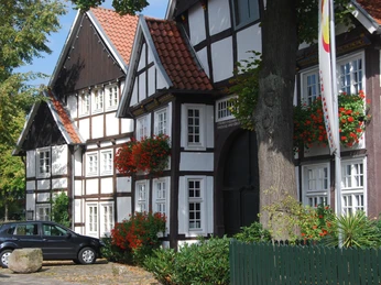 Fachwerkhaus mit roten Dachziegeln, blühenden Geranienkästen und einem schwarzen Auto daneben.