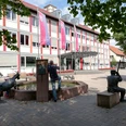 Mensch und Kind betrachten Ziegen am Brunnen vor modernem Gebäude, harmonisch in Lügdes Zentrum.
