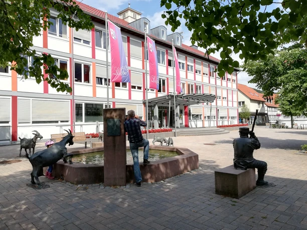 Mensch und Kind betrachten Ziegen am Brunnen vor modernem Gebäude, harmonisch in Lügdes Zentrum.