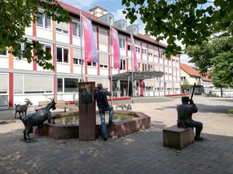 Rathaus Lügde Mensch und Kind betrachten Ziegen am Brunnen vor modernem Gebäude, harmonisch in Lügdes Zentrum.