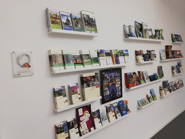 Faltblätter zu regionalen Sehenswürdigkeiten und Aktivitäten auf weißen Regalen an einer Wand.
