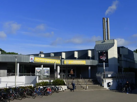 Eingangsbereich der Schwimmoper mit Fahrradstellplätzen, modernem Gebäude und auffälligen Schornsteinen.