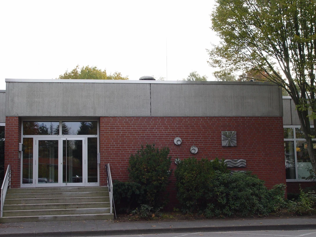 Eingangsbereich eines Gebäudes aus rotem Backstein mit Betonverkleidung und umliegenden Büschen.