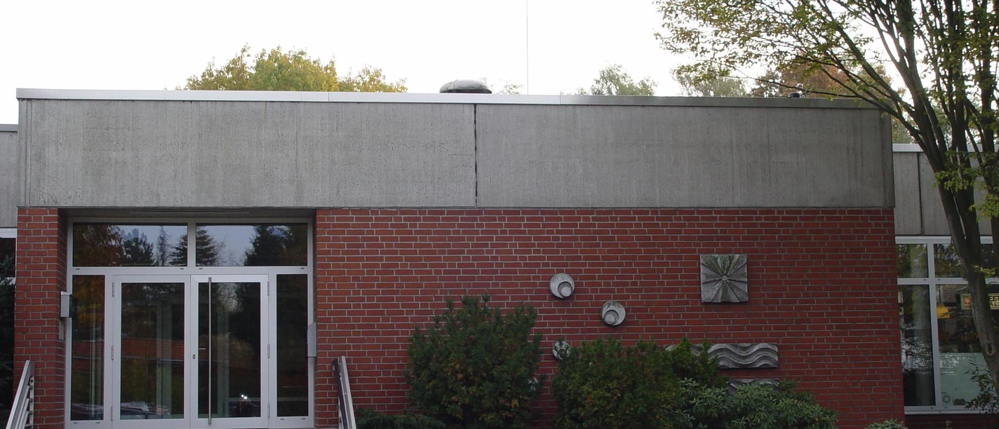 Eingangsbereich eines Gebäudes aus rotem Backstein mit Betonverkleidung und umliegenden Büschen.