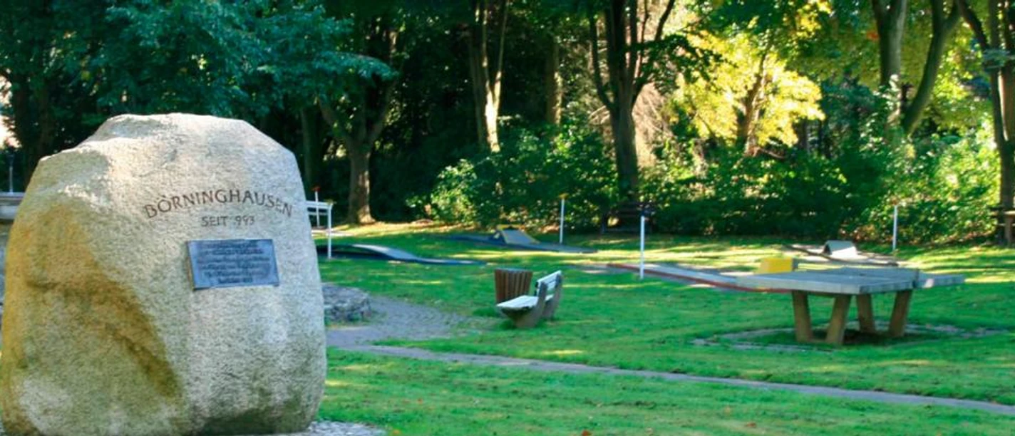 Minigolfanlage im Freizeitpark Eggetal Großer Gedenkstein in grüner Parkanlage mit Minigolfbahnen im Hintergrund, umgeben von Bäumen.