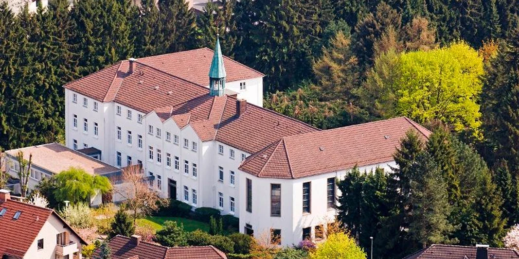 Luftaufnahme des weißen Dreifaltigkeitsklosters, umgeben von dichtem Wald und Wohnhäusern.