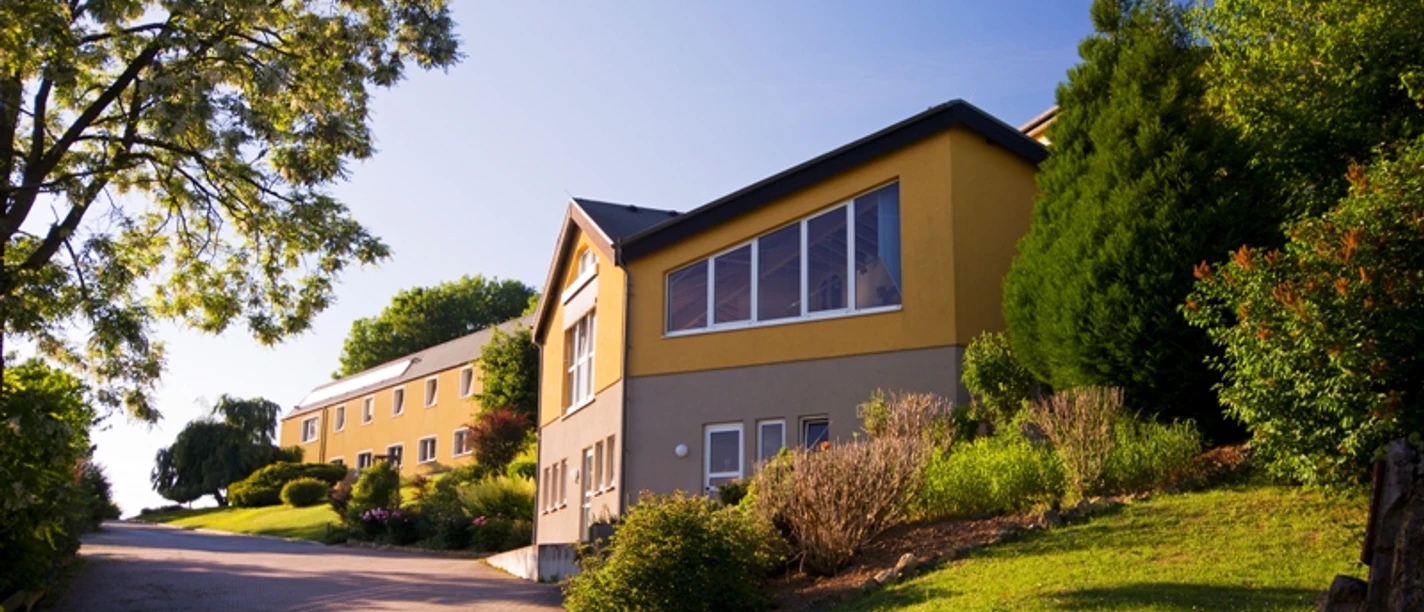 Gelbes Gebäude mit vielen Fenstern auf einem grünen Hügel, umgeben von Bäumen und Sträuchern.