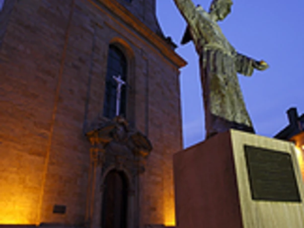Kapuzinerkirche: Statur eines Mönchs vor der beleuchteten Fassade bei Dämmerung. Historisches Bauwerk.