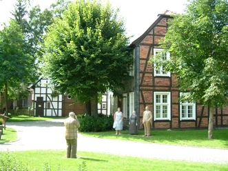 Heimathaus Mastholte Drei lebensgroße Figuren stehen vor historischen Fachwerkhäusern in einer grünen Parkanlage.