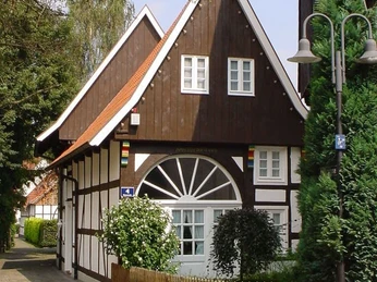 Ein Fachwerkhaus mit weißem Stuck, braunen Holzbalken und roten Dachziegeln, umgeben von Grün.