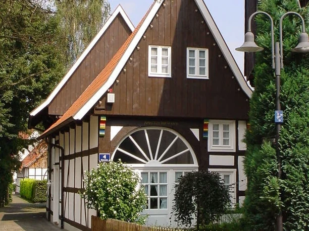 Ältestes Haus in Rietberg Ein Fachwerkhaus mit weißem Stuck, braunen Holzbalken und roten Dachziegeln, umgeben von Grün.