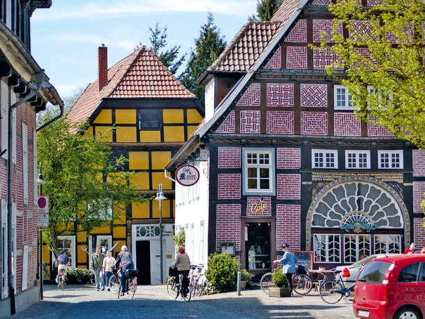 Fachwerkhäuser mit bunten Fassaden an einer gepflasterten Straße; Passanten und Fahrräder davor.