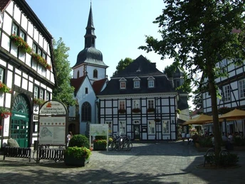 Fachwerkhäuser und Kirche in einer historischen Altstadt mit sonnigem Platz und Café-Tischen.