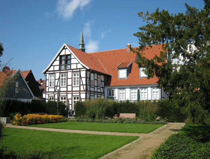 Historisches Fachwerkhaus mit Garten und roten Ziegeldächern, vor blauem Himmel und grünen Büschen.