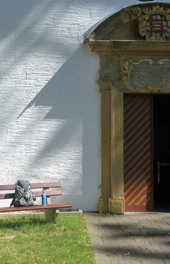 Eingangstor der Kluskapelle St. Lucia umgeben von weißer Wand, Bank und baumbeschattetem Weg.