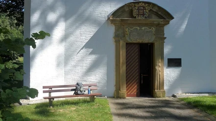 Eingang der Kluskapelle St. Lucia Eingangstor der Kluskapelle St. Lucia umgeben von weißer Wand, Bank und baumbeschattetem Weg.