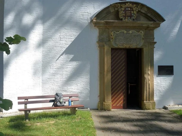 Eingang der Kluskapelle St. Lucia Eingangstor der Kluskapelle St. Lucia umgeben von weißer Wand, Bank und baumbeschattetem Weg.