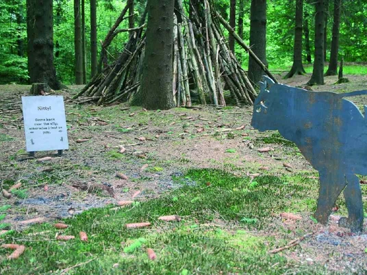Kitty come back Ein hölzernes Kunstwerk im Wald mit Katzenfigur und einem Infobord, umgeben von Bäumen.
