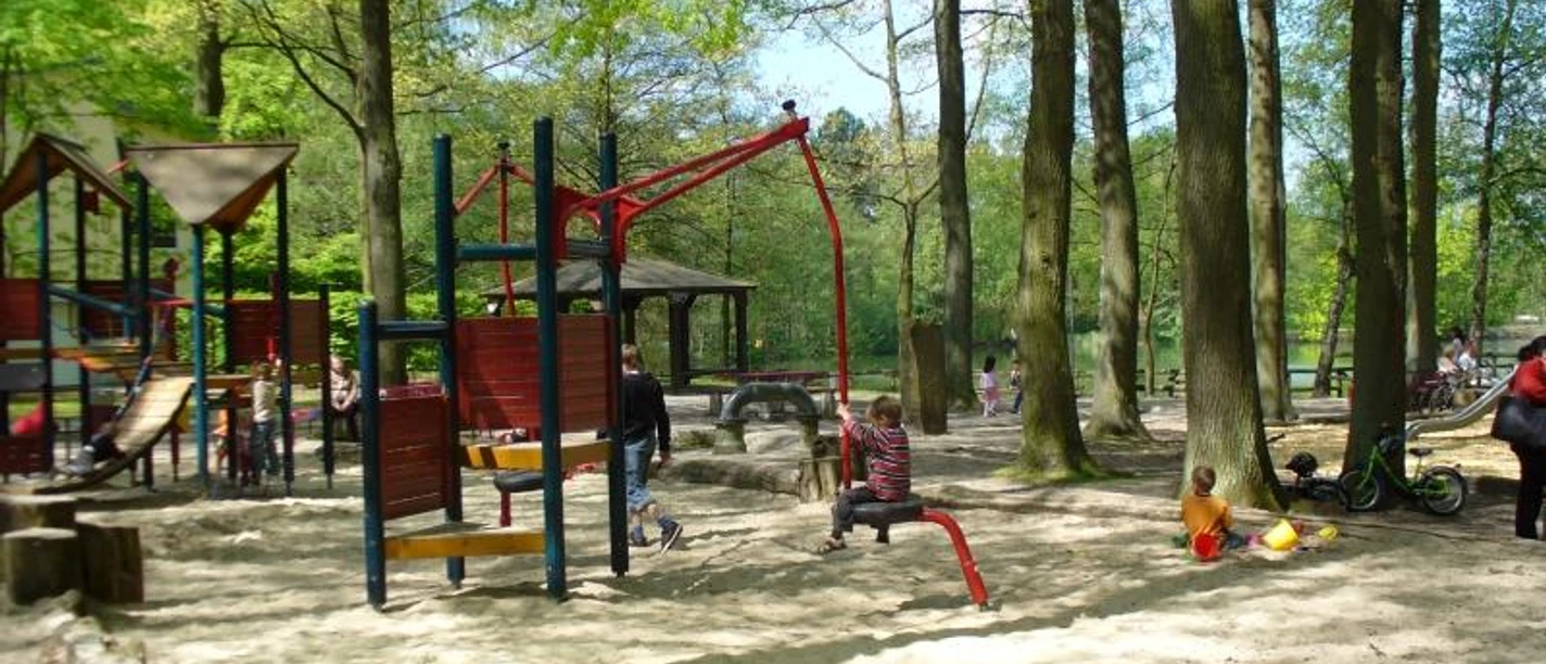 Spielplatz bei den Fischteichen Spielplatz mit Holzspielgeräten, Schaukeln und sandigem Boden, umgeben von hohen Bäumen.